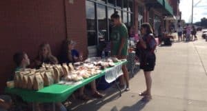 A downtown sidewalk with people shopping at a table full of baked goods